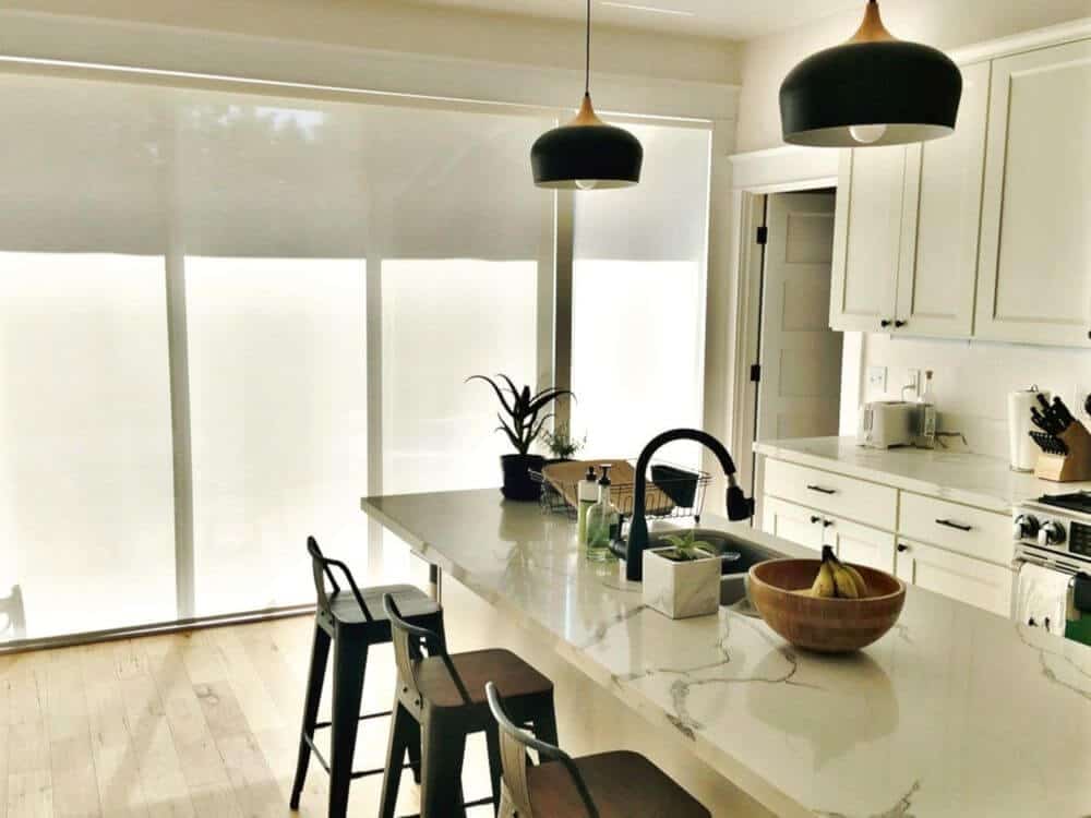 这间厨房设有一个狭窄的中央岛台，带大理石台面，早餐吧台由吊灯照明。该区域的特色是带遮阳板的窗户。