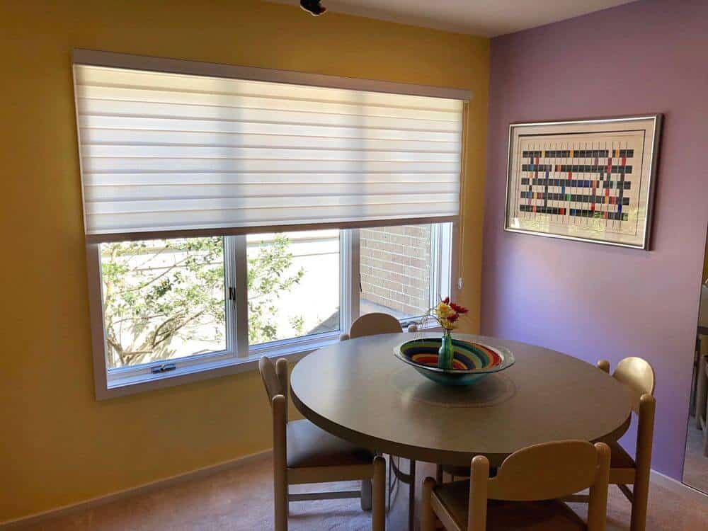 一个简单的用餐区，有一个棕色圆形的四人用餐角，周围是黄色和紫色的墙壁。餐桌设置在有过渡窗帘的玻璃窗附近。
