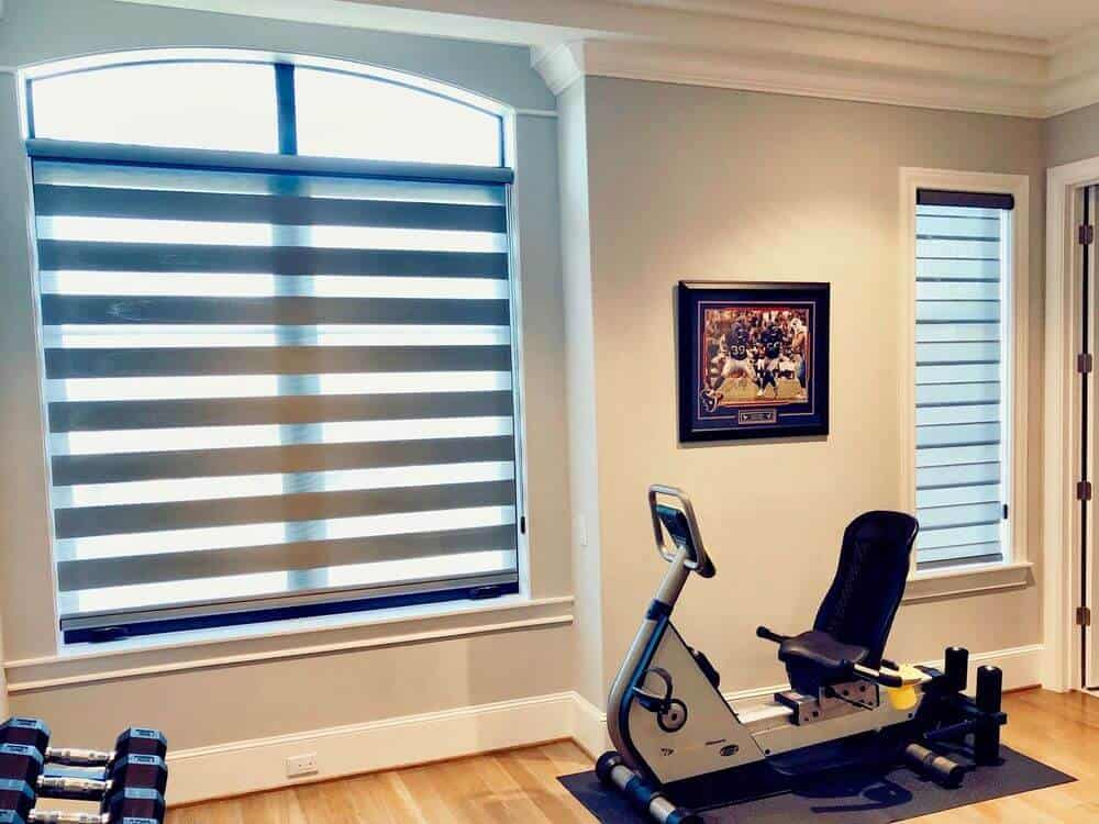 一个家庭健身房，有浅灰色的墙壁和硬木地板，以及白色的天花板。房间还设有带有过渡阴影的窗户。