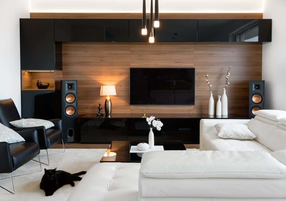 平均大小的现代客厅，具有高光泽的凹进式橱柜，黑色皮革扶手椅，白色l形沙发，壁式电视和光滑的环境照明。