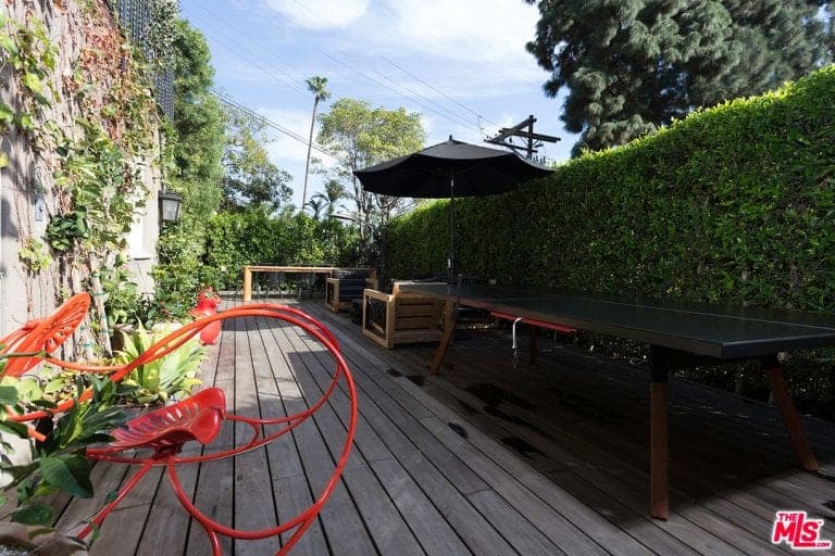 一个独特的红色躺椅在这个放松的甲板上脱颖而出，上面有一张长桌和一把独立的伞下的木椅。它被一株树篱和覆盖着匍匐植物的混凝土栅栏夹在中间。