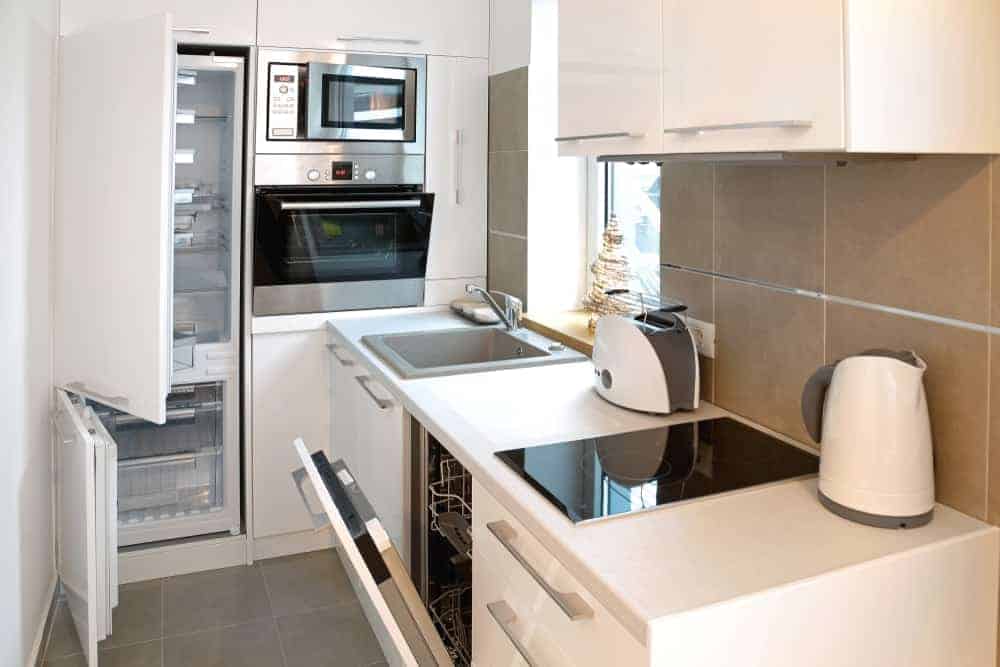 一个现代化的白色小厨房，配有现代化的电器，土黄色的后挡板砖和灰色的地砖。