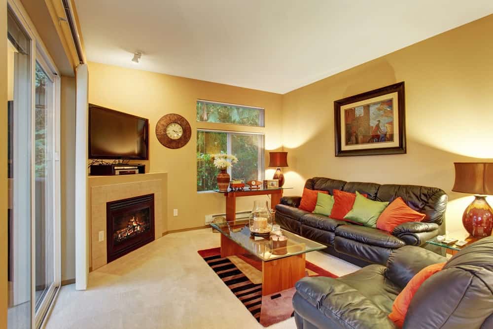 中等大小的客厅，黄色的墙壁，米色的地毯地板，角落壁炉和电视，皮革座椅，彩色枕头，玻璃顶桌，挂钟和一件可爱的艺术品。