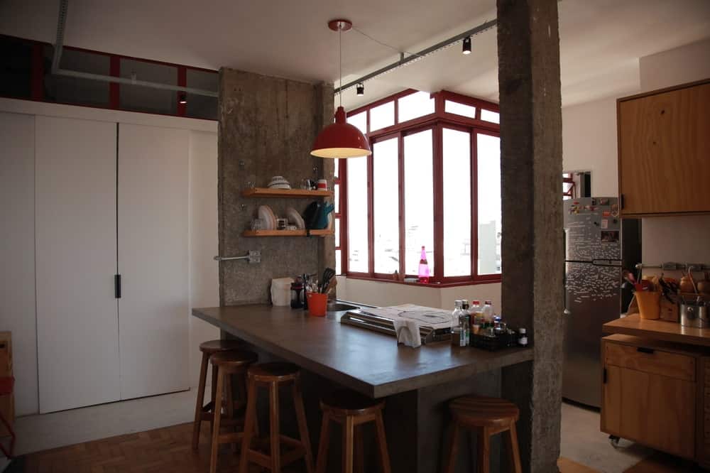 这是厨房，有一个大的混凝土厨房岛连接到两根柱子上，并在两侧搭配早餐吧台的凳子。这些由吊灯和远侧的一排角窗补充。