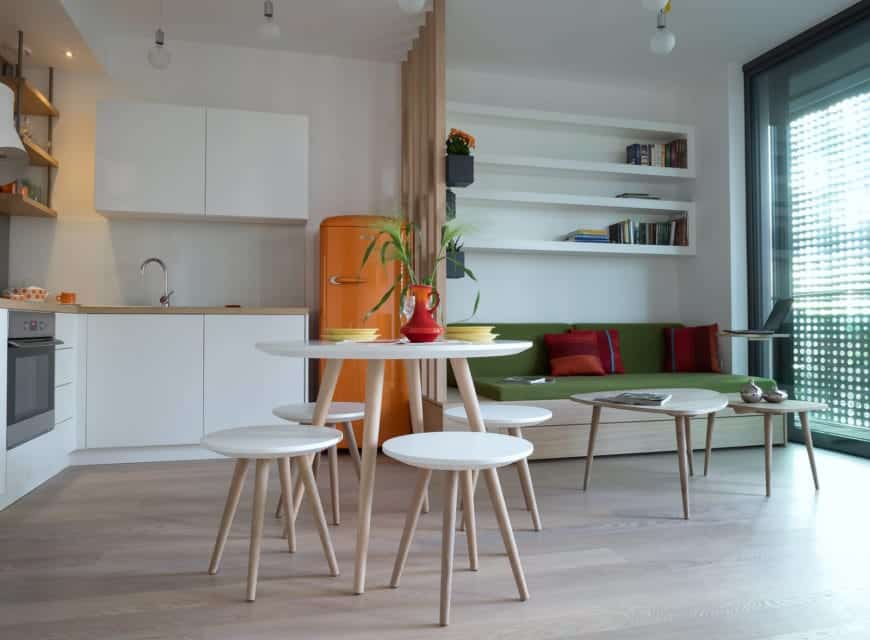 就餐厨房与生活空间融为一体。它展示了一套圆形的餐具和一个大胆的橙色冰箱，在白色的橱柜和墙壁的映衬下很显眼。