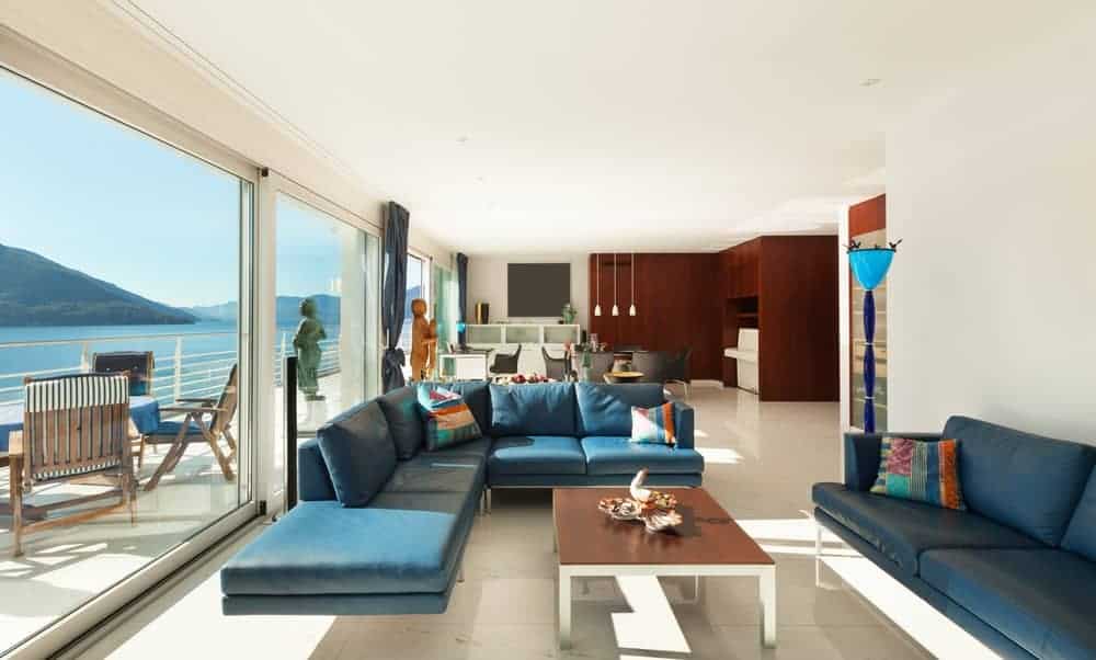 的客厅拥有蓝绿色截面与彩色枕头和一个木质咖啡桌在大理石瓷砖地板。它有白墙和大玻璃窗的海景。