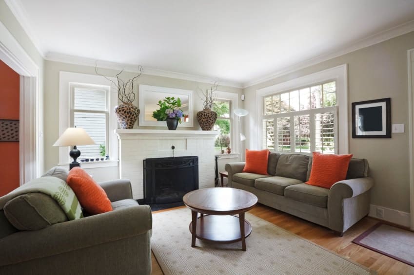 橙色抱枕为客厅的灰色区域增添了不错的口音，中性色调的浅灰色墙壁和白色壁炉。中间有一张圆形的咖啡桌，放在有图案的地毯上。