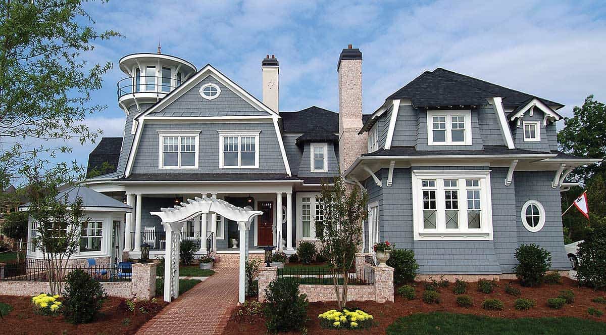 这是一个近距离观察的木瓦风格的房子的正面外观，灰色的外墙，白色的窗户和门，以及高高的砖烟囱，与前面草坪的栅栏相匹配。