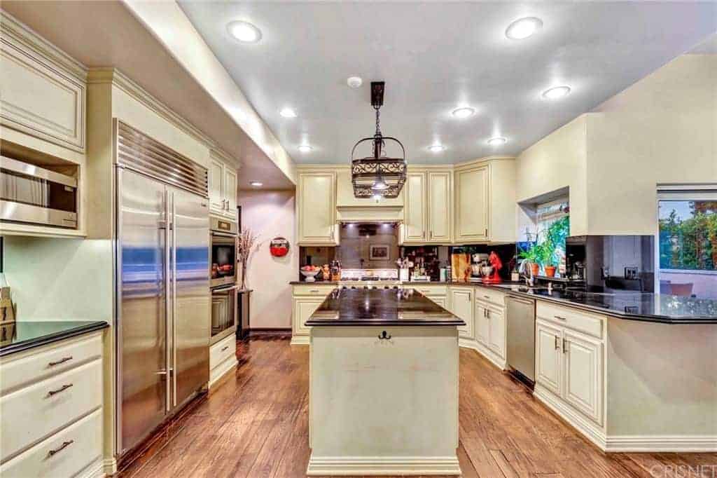 这个厨房的米色橱柜和抽屉有金属把手，与厨房岛台的黑色窗台上的金属悬垂照明设备相协调，与l形半岛相匹配。