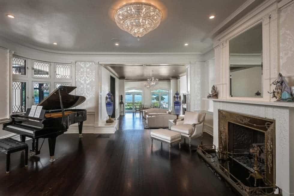 这是一个大房间，在远端设有一个客厅，在大型壁炉旁边有一把宏伟的钢琴，上面有一个白色的地幔，在宽阔的豪华深色硬木地板上脱颖而出，上面放着水晶冲洗式照明。图像由toptenrealestatedeals.com提供。