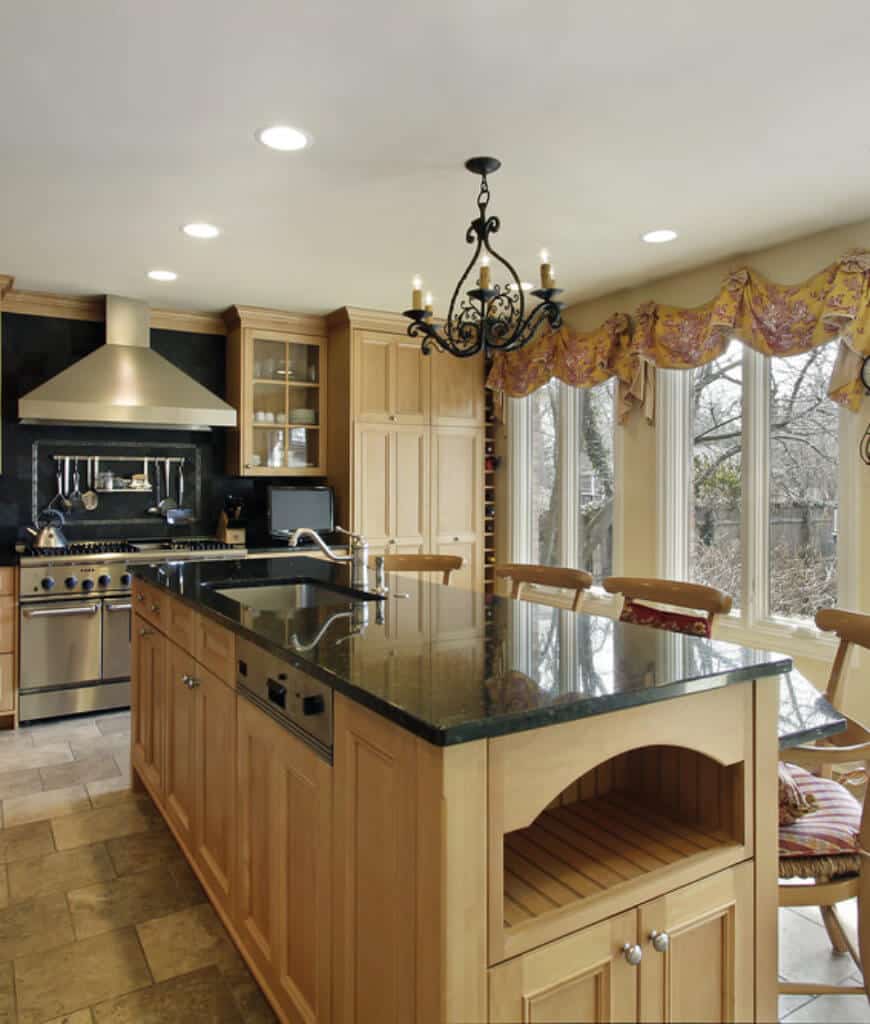 一盏锻铁枝形吊灯悬挂在这间厨房里的轻木早餐吧台上，厨房的地面是石灰石，玻璃框窗户上装饰着迷人的黄色镶边。