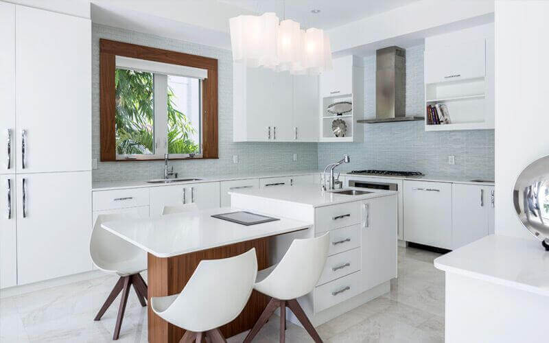 这个白色厨房有一个白色的中央岛台，内置白色桌面，配有现代椅子，并配有令人惊叹的吊灯。