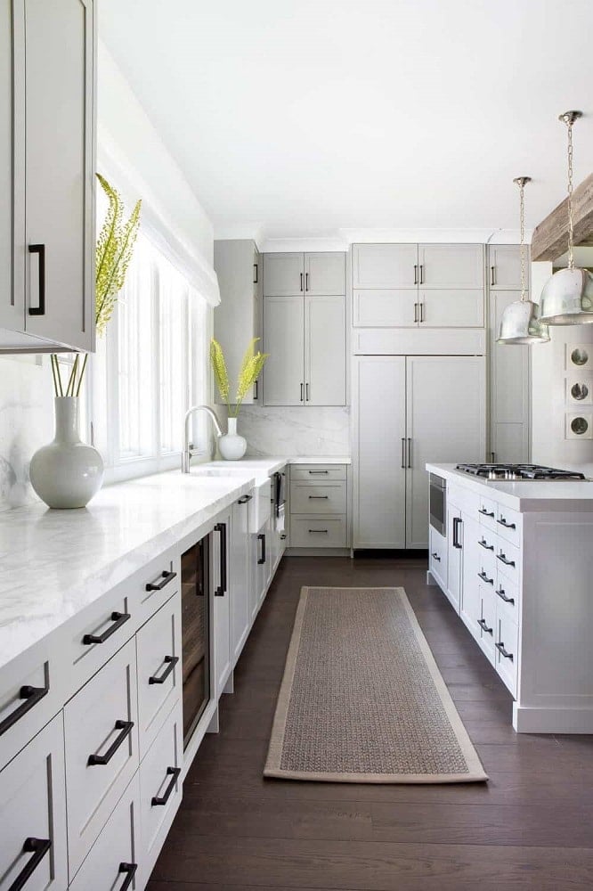 这个狭长的厨房的黑色把手和固定装置与明亮的白色橱柜形成鲜明对比，也与深色硬木地板形成对比。水龙头上方的窗户使这些区域更加明亮。