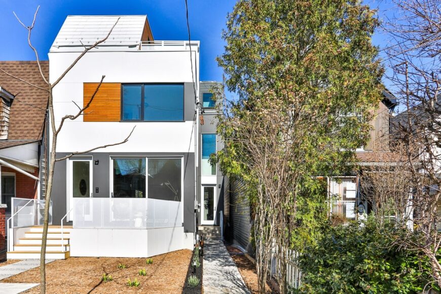 这个现代家用的家居拥有白色和灰色的外墙，侧面的大型玻璃窗装饰着木板。在这里，您还可以通过长途狭窄的走道看到侧入口。