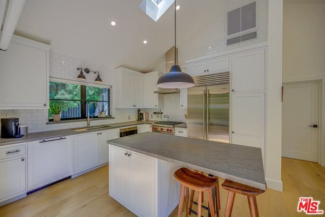 这是一个简单而迷人的厨房，白色的拱形天花板，白色的墙壁和白色的橱柜与厨房岛台相匹配，厨房岛台的灰色台面上有一张嵌壁式桌子。