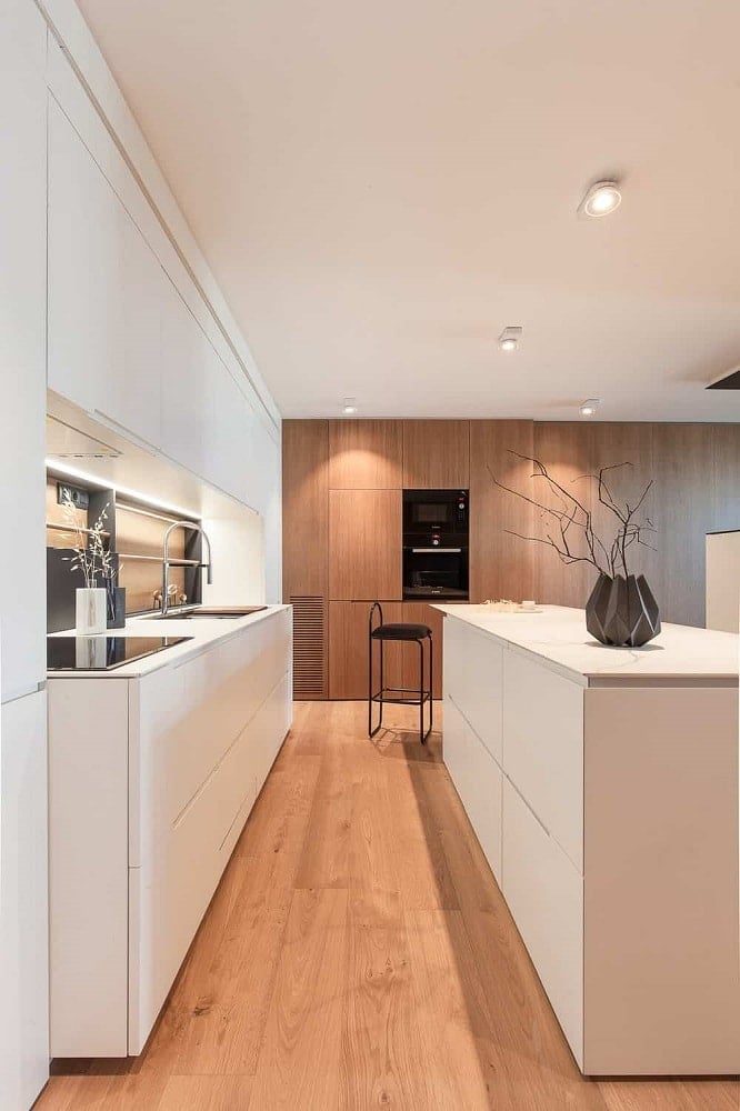 这是现代化的狭长厨房，白色的现代橱柜与白色的台面和白色的天花板相匹配。这与硬木地板和远端墙壁的木质色调相辅相成。