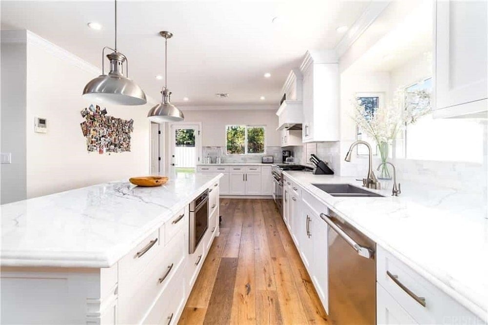 厨房岛台的白色台面上安装了几盏不锈钢吊灯，与白色橱柜中放置的现代电器相匹配，在这个狭长的厨房中，硬木地板与之相辅相成。