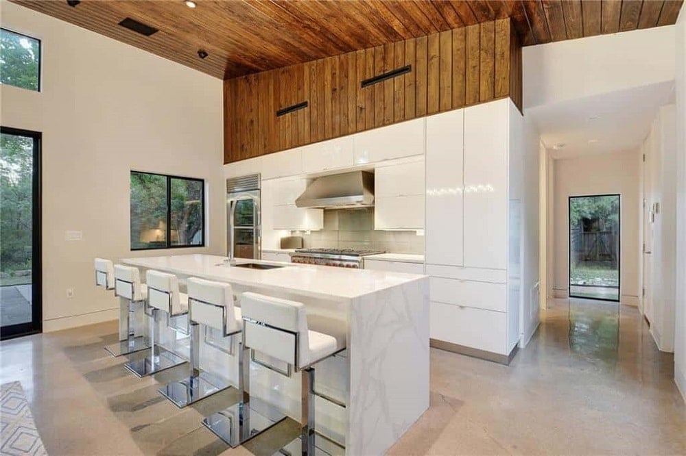 狭长的厨房里现代化的白色橱柜与早餐吧的白色台面和白色软垫凳子完美搭配。硬木地板和木制吊顶作为补充。