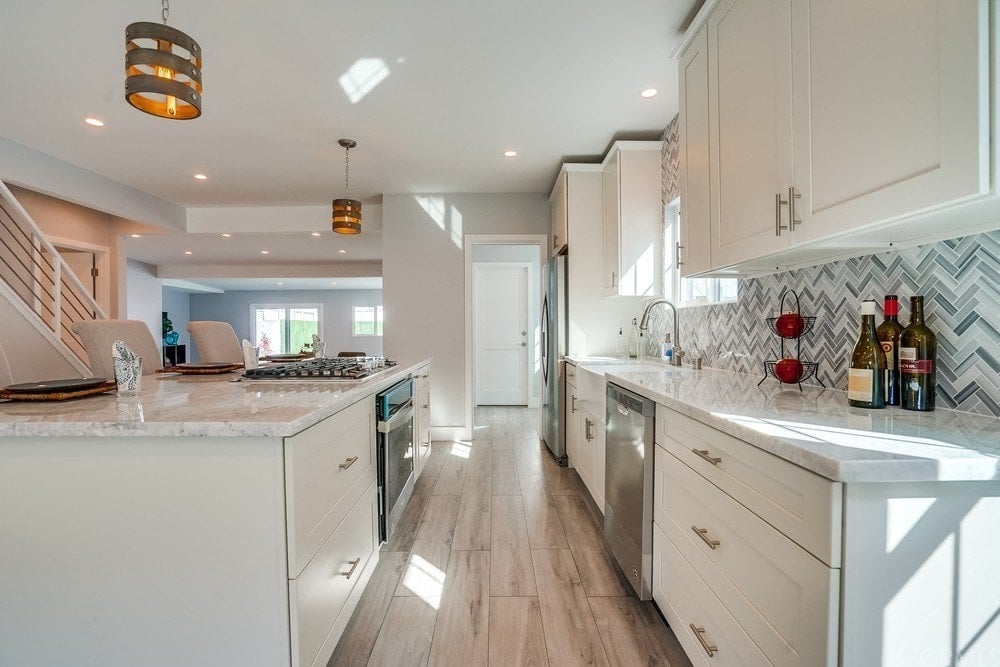 这是一个简单而优雅的厨房，经典的白色橱柜用来放置不锈钢用具，辅以硬木地板和厨房岛上的装饰性吊灯。