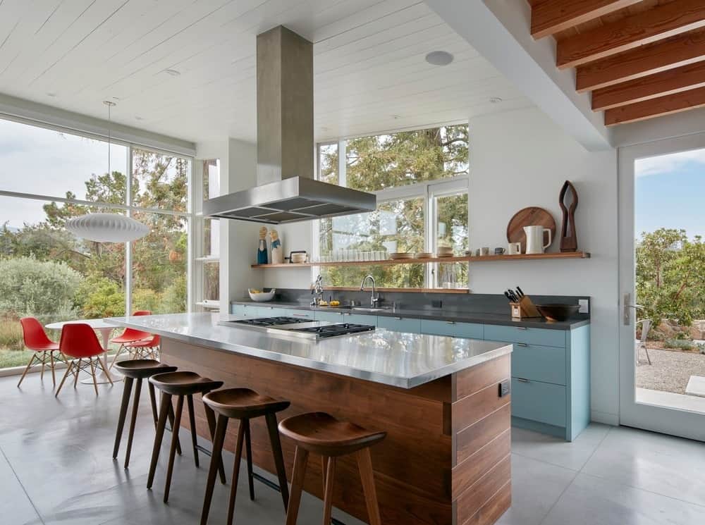 这间厨房有一个长长的厨房岛台，配有供早餐吧台使用的木凳。它的顶部有一个不锈钢通风口，通风口来自高高的白色棚顶。