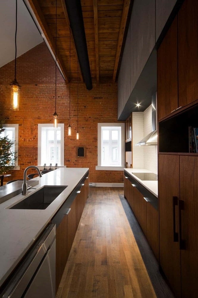 这是一个狭长的厨房，配有可爱的深色硬木地板，与两侧的木制橱柜相匹配，远处还有一面大红砖墙。