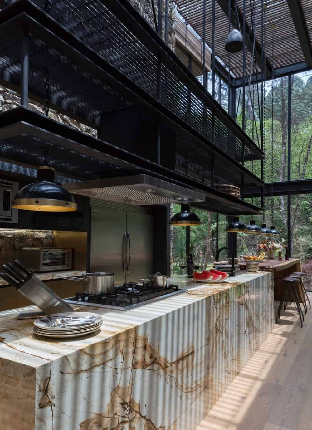 这是一间狭长的厨房，有一个巨大的大理石瀑布式厨房岛台，顶部有金属梯级，远端有一张嵌壁式木桌，配有木凳。