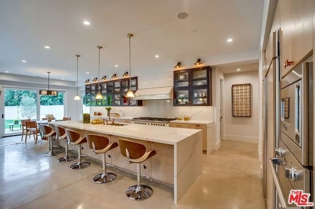 这个狭长的厨房配备了现代化的白色瀑布台面，与后挡板和天花板相匹配，天花板上有厨房岛台上方的吊灯。