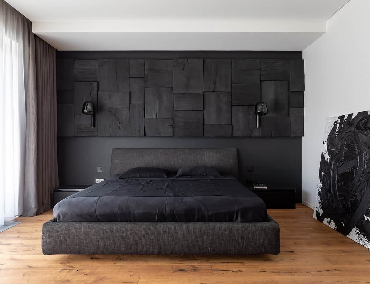 这间漂亮的卧室里有一张黑色的床和一个黑色的床头板，与后面有纹理的黑色墙壁相匹配。硬木地板、明亮的白色天花板和侧面的墙壁作为补充。