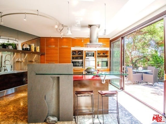 这是一个可爱的厨房，明亮的白色天花板，烹饪区上方有一个不锈钢通风口。这个视图还显示了厨房岛边的内置玻璃桌子和几把凳子。