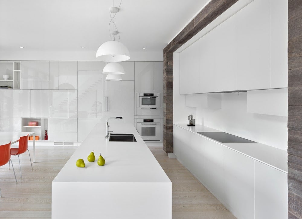 这是一个美丽的白色厨房，它的现代白色橱柜和白色厨房岛的狭窄和长设计。浅色的硬木地板和厨房岛台上方的白色吊灯与之相辅相成。