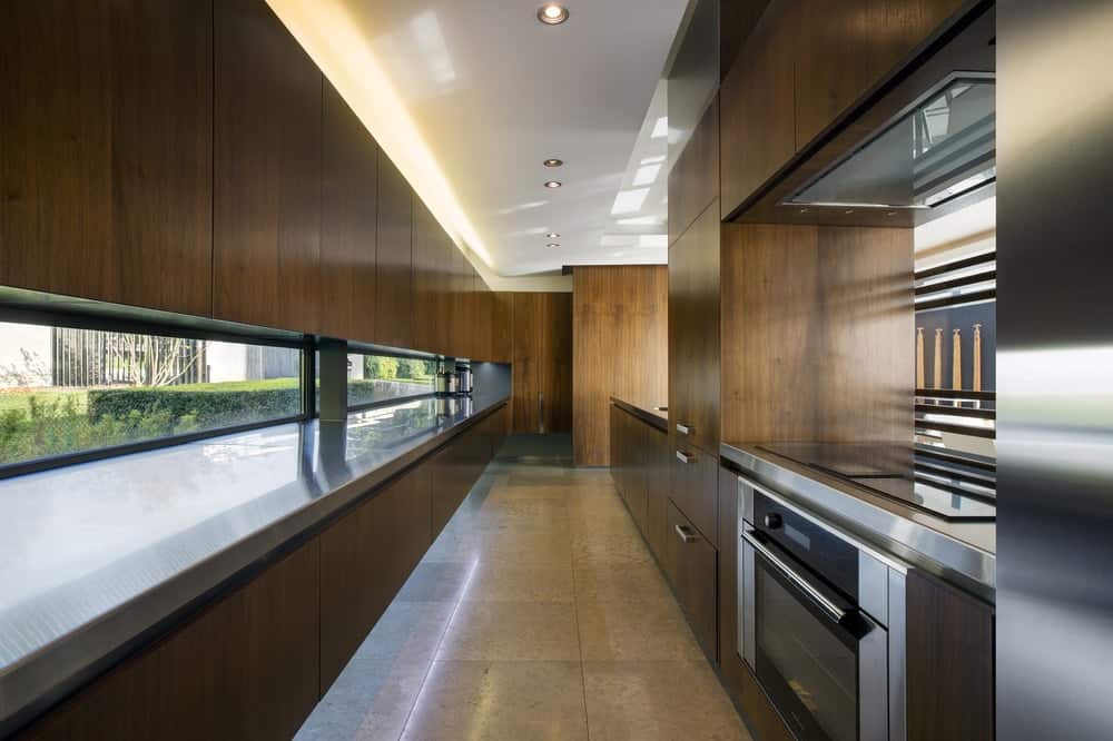 这是一个狭长的厨房，配有深棕色木制现代橱柜，用于放置现代不锈钢用具。天花板和一排窗户补充了这些黑暗元素。