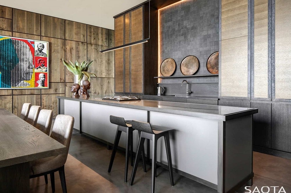 这是一个精致而豪华的狭长厨房，宽敞的厨房岛台配有光滑的深灰色台面，用餐区旁边的早餐吧配有深色凳子。