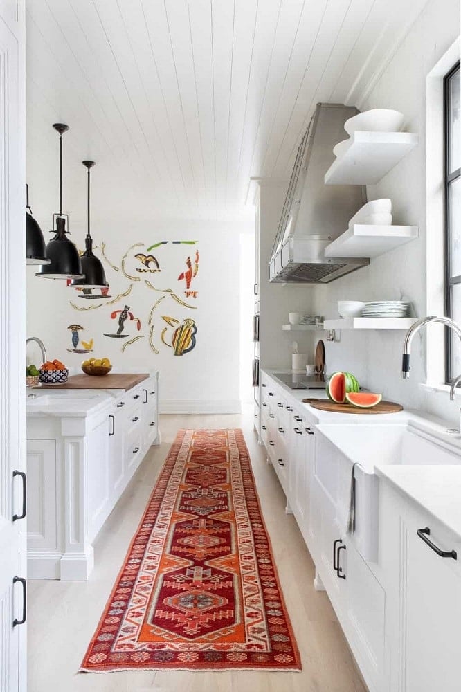 这间厨房狭长的白色地板上装饰着彩色图案的区域地毯，与明亮的白色橱柜和台面形成鲜明对比。厨房岛台的顶部是一排黑色吊灯。