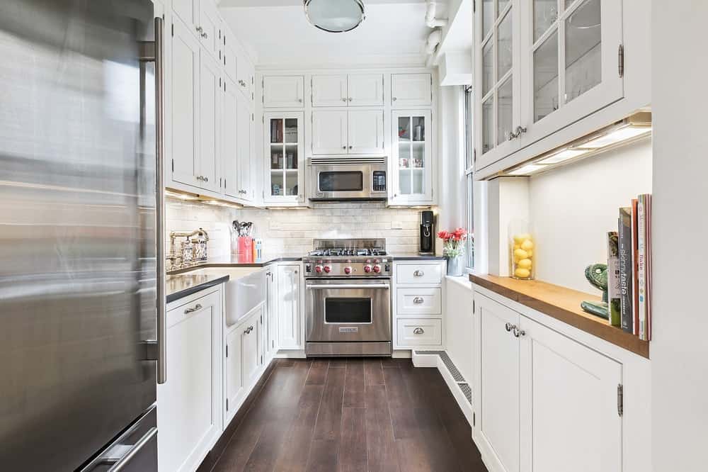 这是公寓的厨房风格，有丰富的深色硬木地板，与白色的摇床柜和抽屉形成对比。在最远的一端是不锈钢炉顶烤箱。