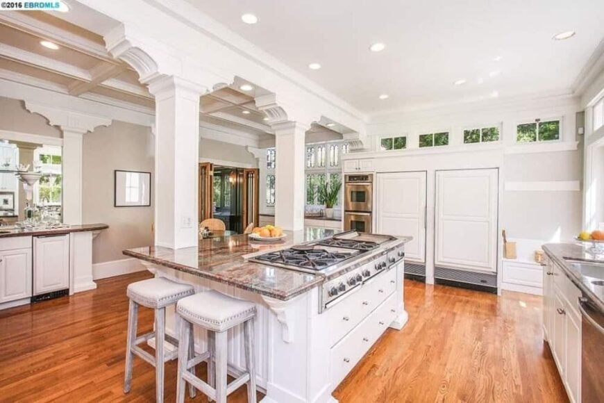 这个美丽的厨房有明亮的白色橱柜，与白色的大厨房岛相匹配。它在硬木地板的衬托下格外显眼，并通过几根带拱门的白色柱子与白色天花板相连。