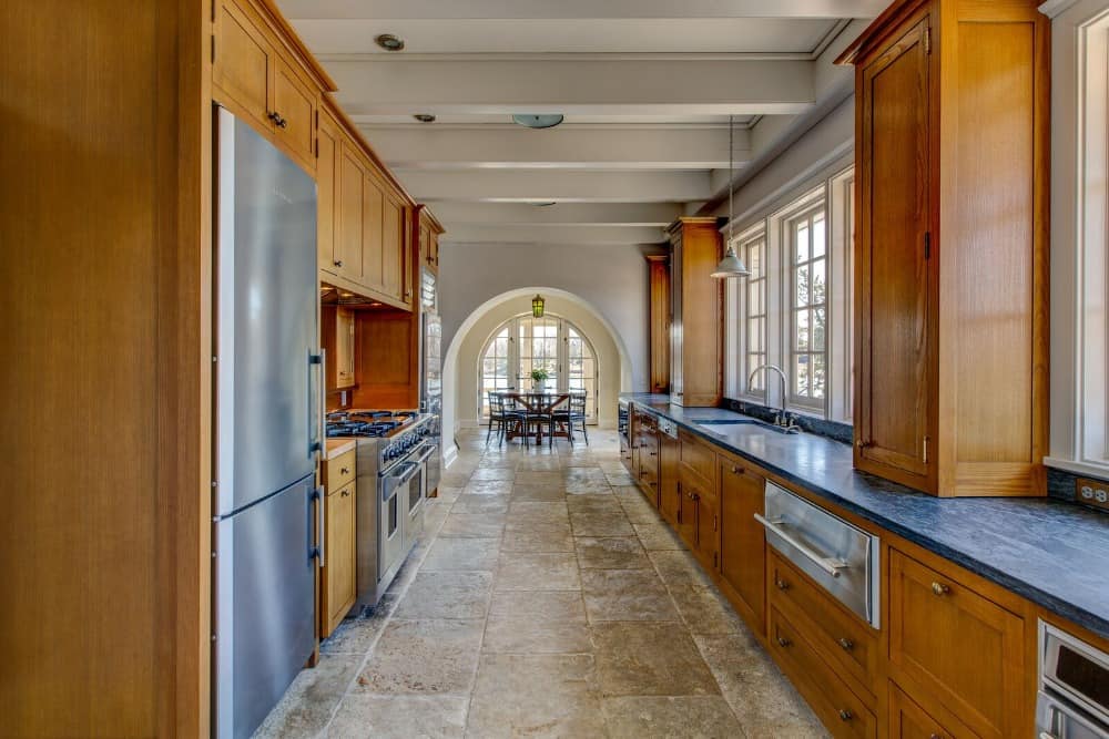 这是一个可爱的狭长厨房，配有时尚的棕色橱柜，里面装有不锈钢用具，辅以米色的地板和横梁天花板。