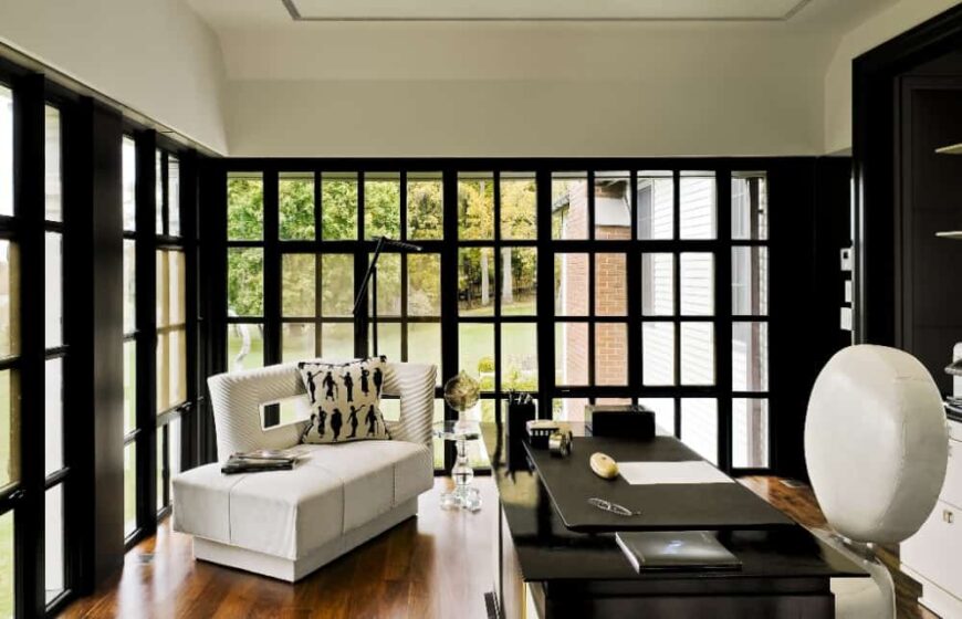 这是一个时尚的家庭办公室，有一张光滑的黑色桌子，与周围的窗框形成对比，白色的天花板和角落里的现代沙发形成对比。