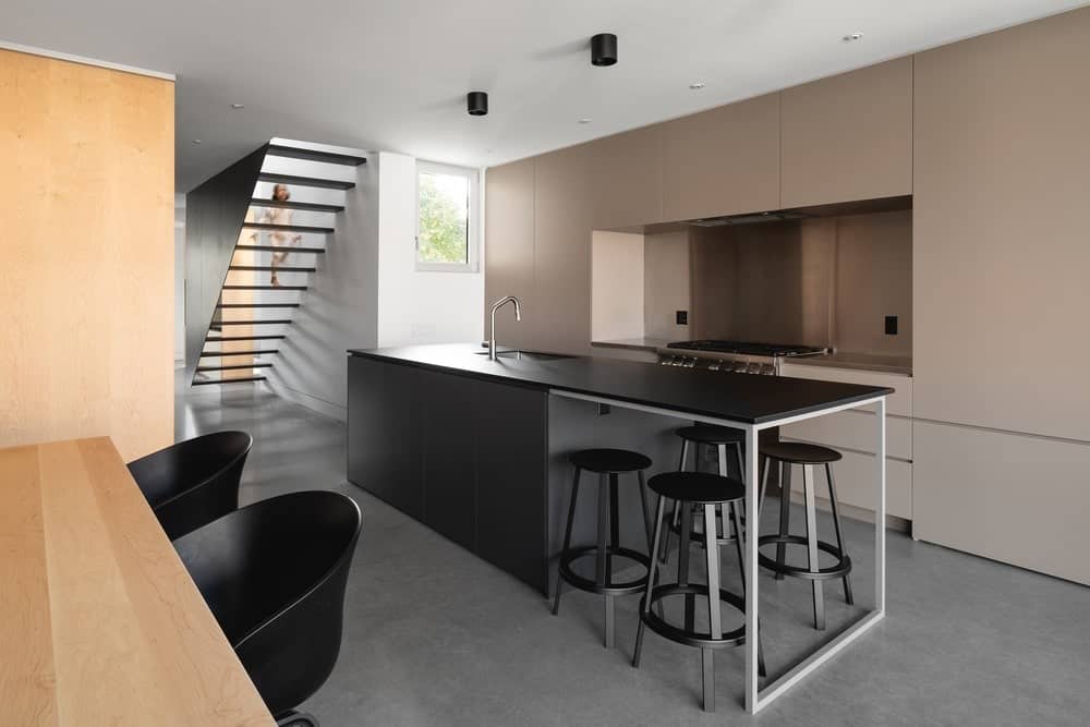 这是一个现代厨房，有一个长长的黑色厨房岛台，使它的设计又长又窄。岛的边缘延伸到一张配有黑色凳子的现代嵌入式桌子。
