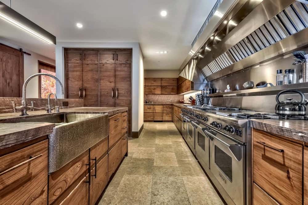 这个华丽的长而窄的厨房有深棕色的橱柜，辅以现代不锈钢电器，顶部是明亮的白色天花板，带有嵌入式灯。