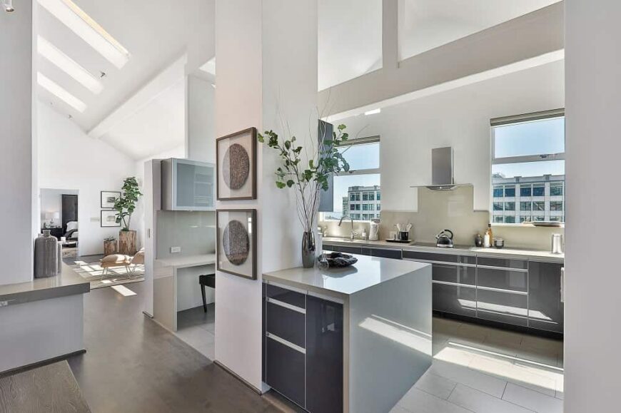 这间现代厨房配备了现代的深灰色橱柜，与带有灰色大理石瀑布台面的小厨房岛相匹配。与之形成对比的是连接到高高的白色天花板的白色柱子。