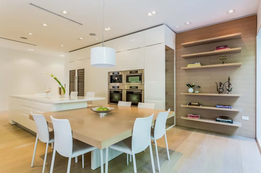 这是明亮的厨房，白色的橱柜与墙壁和天花板相匹配。大厨房岛台有一张嵌壁式木桌，搭配白色现代椅子，顶部有一盏白色吊灯。