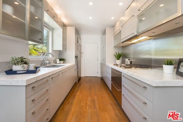 这是一个狭长的厨房，现代化的橱柜色调轻盈，里面摆放着不锈钢用具。丰富的硬木地板和明亮的白色天花板与凹形灯相辅相成。