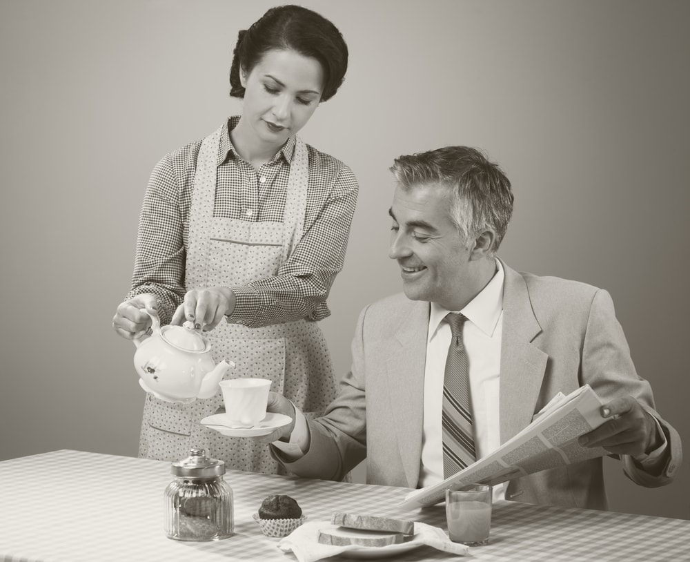 1950年代的妻子穿着围裙,倒茶给丈夫当他坐在桌子上吃早餐和报纸。