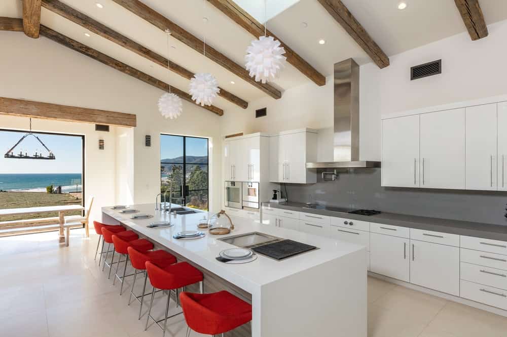 这个明亮的白色厨房有一个长长的白色瀑布厨房岛台，搭配对比鲜明的红色凳子。隔着这片区域就是带有深灰色后挡板的烹饪区。