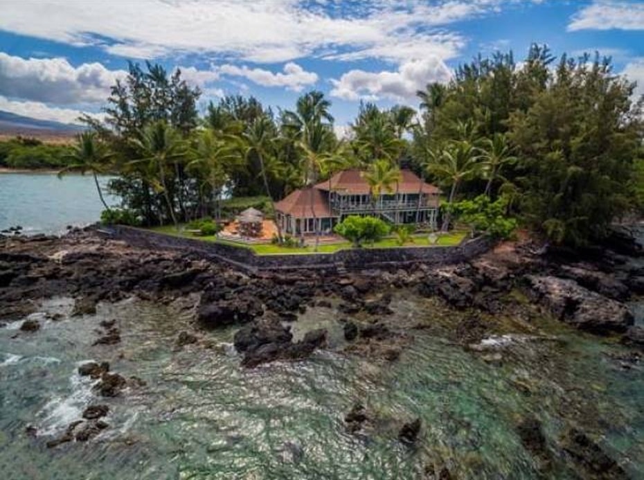 这是海滩别墅的航拍照片，它在岩石边缘面朝大海。你可以在这里看到，房子有赤陶土色调的屋顶，使它在高大的热带树木的背景下脱颖而出。图片来自Toptenrealestatedeals.com。
