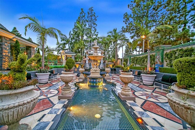 这是游泳池的景观，它的走道上有图案瓷砖，装饰着基座花盆，中间有一个大喷泉。然后这些被背景中的高大树木所平衡。图片来自Toptenrealestatedeals.com。