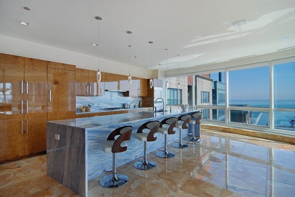 这是一间厨房，有一个白色大理石瀑布台面，搭配现代的早餐吧台凳子。图片来自Toptenrealestatedeals.com。