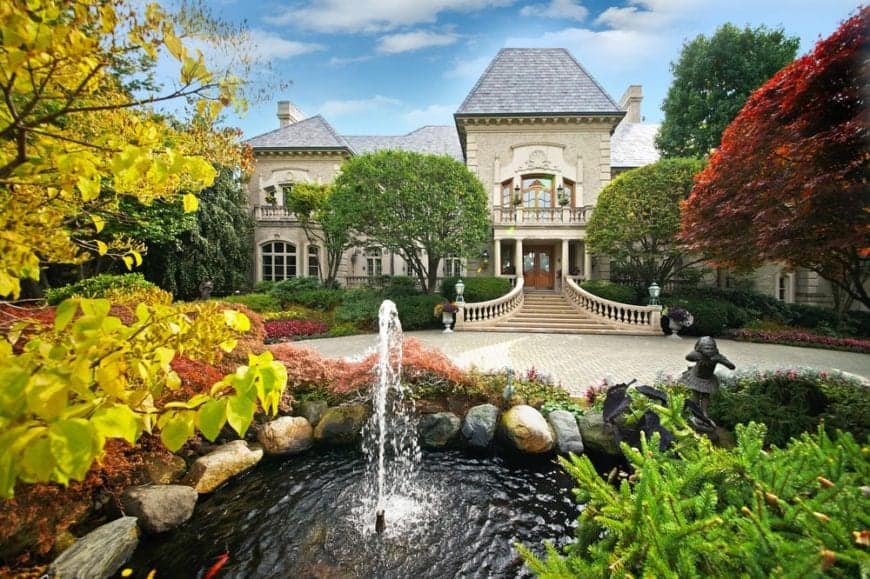 这是房屋正面的鸟瞰图，与周围高大的树木、五颜六色的灌木和一个带喷泉的池塘景观相辅相成，池塘对面是主入口的大庭院。