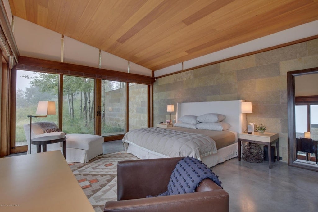 这是一间卧室，天花板是木棚，床是浅灰色的，床头板有软垫，两侧有台灯。图片来自Toptenrealestatedeals.com。
