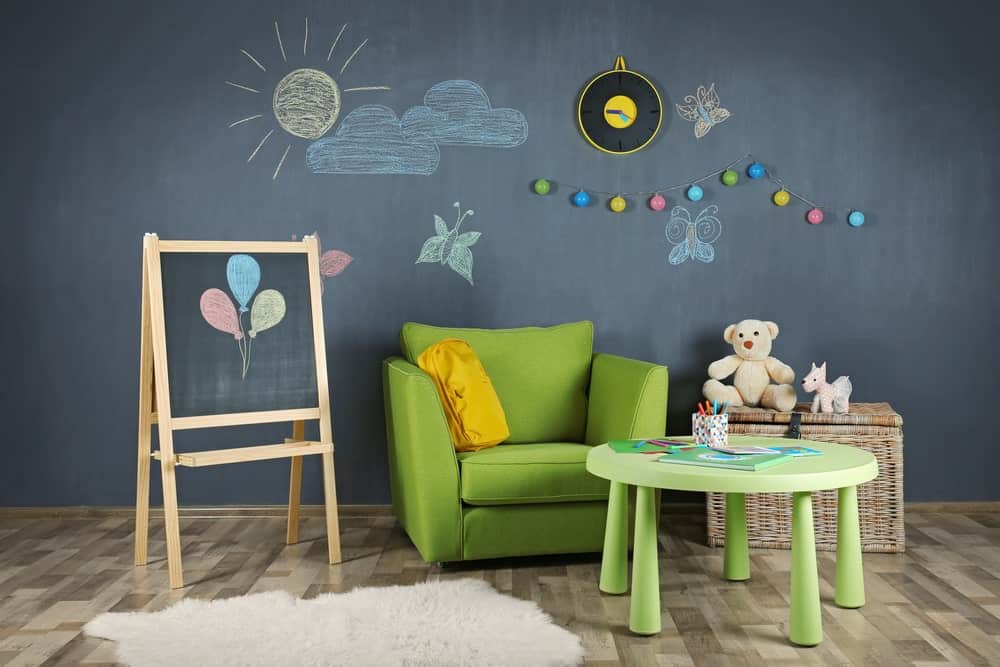 儿童房有绿色扶手椅，圆桌，硬木地板，藤条玩具储物柜，一面黑板墙。